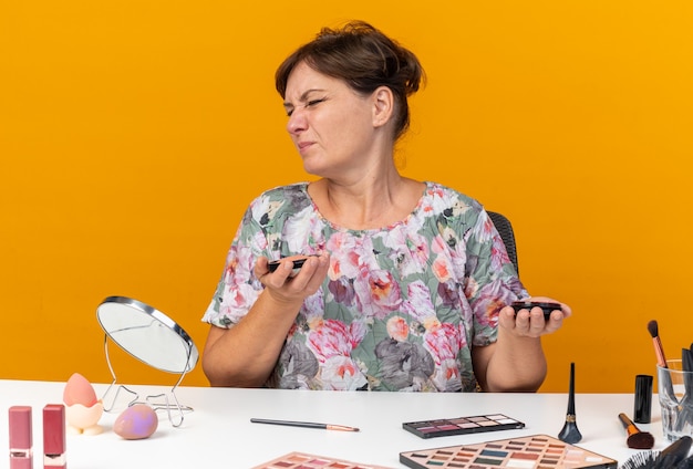 Mujer caucásica adulta disgustada sentada a la mesa con herramientas de maquillaje sosteniendo rubor aislado en la pared naranja con espacio de copia