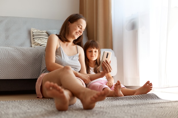 Mujer caucásica adulta joven positiva con ropa de estilo casual posando interior en la sala de estar con su hija mientras está sentada en el piso cerca del sofá y la ventana, viendo videos divertidos con el niño.