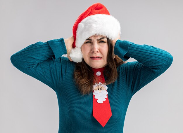 Mujer caucásica adulta molesta con gorro de Papá Noel y corbata de santa cubriendo las orejas con las manos aisladas sobre fondo blanco con espacio de copia