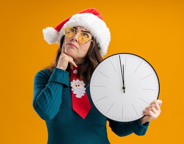 Mujer caucásica adulta pensativa en gafas de sol con gorro de Papá Noel y corbata de santa sostiene el reloj y pone el dedo en la barbilla mirando hacia arriba aislado en la pared naranja con espacio de copia