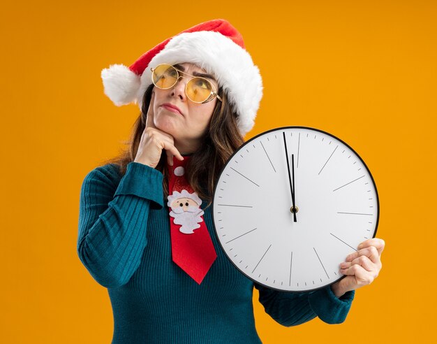 Mujer caucásica adulta pensativa en gafas de sol con gorro de Papá Noel y corbata de santa sostiene el reloj y pone el dedo en la barbilla mirando hacia arriba aislado en la pared naranja con espacio de copia