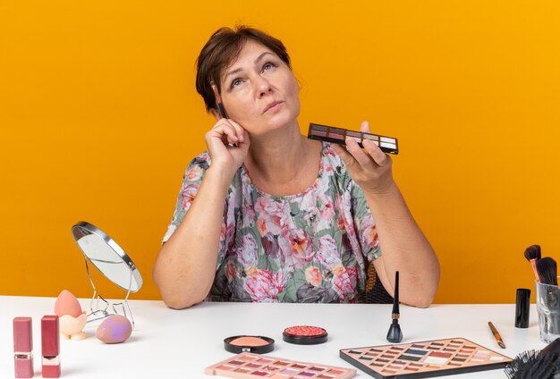 Mujer caucásica adulta pensativa sentada a la mesa con herramientas de maquillaje sosteniendo la paleta de sombra de ojos y pincel de maquillaje mirando hacia arriba