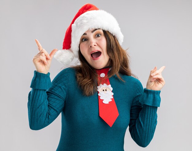 Mujer caucásica adulta sorprendida con gorro de Papá Noel y corbata de santa apuntando hacia arriba aislado sobre fondo blanco con espacio de copia