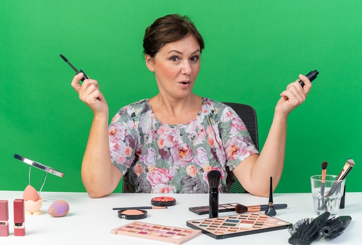 Foto gratuito mujer caucásica adulta sorprendida sentada en la mesa con herramientas de maquillaje con rímel