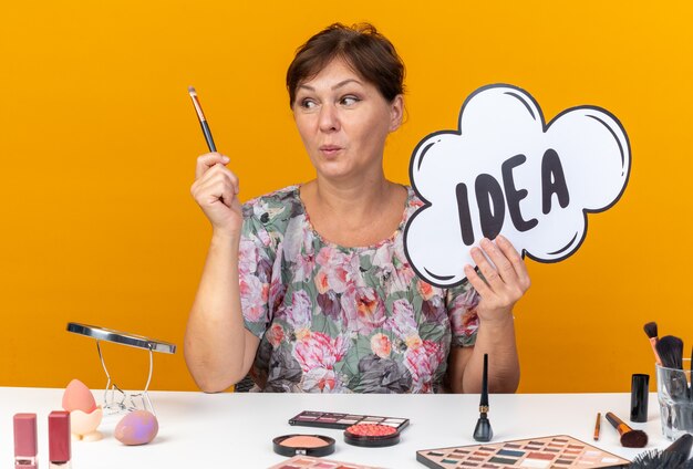 Mujer caucásica adulta sorprendida sentada en la mesa con herramientas de maquillaje sosteniendo la burbuja de la idea y mirando el pincel de maquillaje