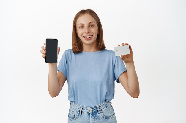 Foto gratuita mujer caucásica alegre que muestra la pantalla vacía de su teléfono y su tarjeta de crédito, sonriendo feliz, recomendando un banco, una aplicación para rastrear las finanzas, de pie contra un fondo blanco.