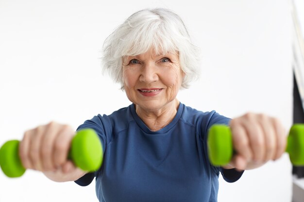 Foto gratuita mujer caucásica anciana feliz enérgica activa con cabello gris disfrutando de ejercicios físicos en el interior, entrenando en casa usando pesas, sonriendo ampliamente. enfoque selectivo en el rostro de la mujer.