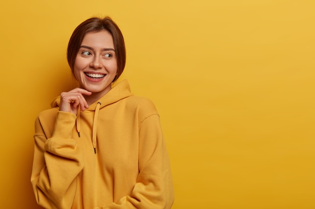 Una mujer caucásica de aspecto agradable mantiene la mano en la cara, mira hacia otro lado con satisfacción, nota algo agradable, usa una sudadera con capucha informal, aislada en la pared amarilla, espacio en blanco a un lado para su promoción