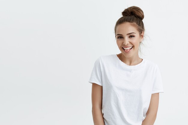 Mujer caucásica atractiva positiva con bollo, sacando la lengua y sonriendo, mirando contento y complacido