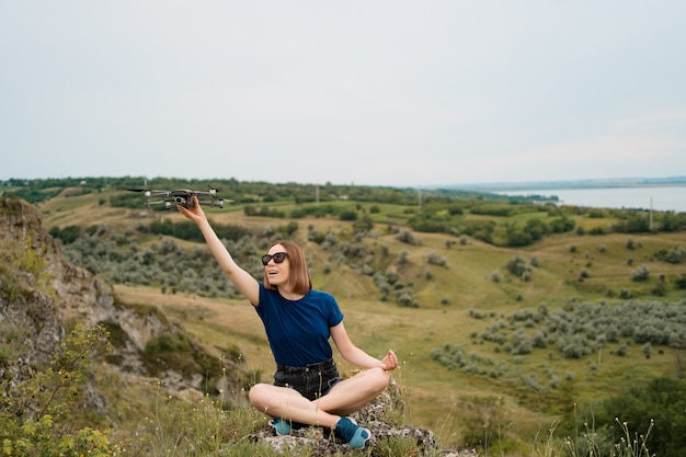 Una mujer caucásica con un avión no tripulado en la mano, sentada en una colina rocosa verde con cielo