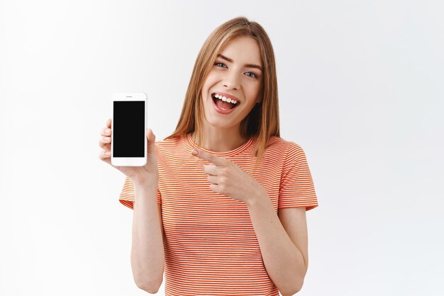 Mujer caucásica bastante alegre con camiseta a rayas sosteniendo un teléfono inteligente apuntando a la pantalla del teléfono móvil negro y sonriendo mientras discute la nueva aplicación recomienda seguir su fondo blanco en las redes sociales