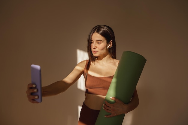 Mujer caucásica bastante joven con colchoneta de fitness toma selfie en iphone en interiores Mujer morena en ropa deportiva disfruta de la tecnología moderna Concepto estilo de vida ocio