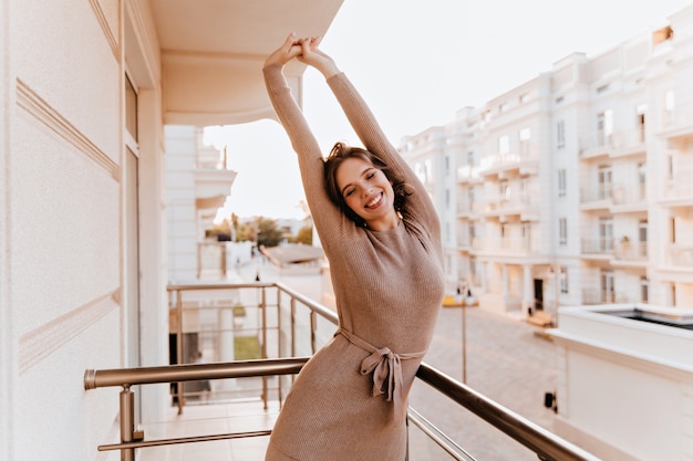 Mujer caucásica bien formada en vestido marrón que se extiende en el balcón. Chica morena de ensueño disfrutando de la mañana en la terraza.