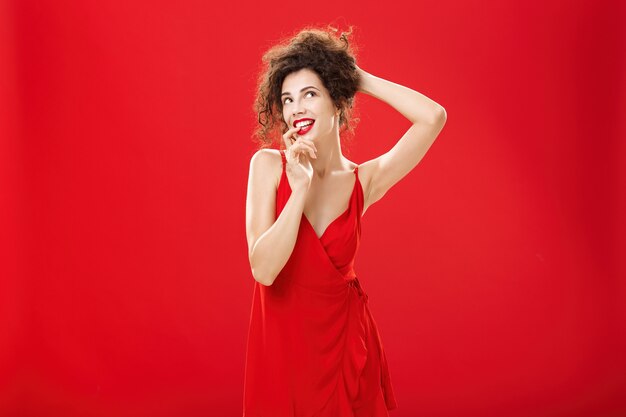 Mujer caucásica de buen aspecto soñadora y elegante en elegante vestido de fiesta peinando el cabello sosteniendo mechones rizados con la mano mordiendo el dedo coqueta mirando en la esquina superior derecha nostálgica, soñando despierto.