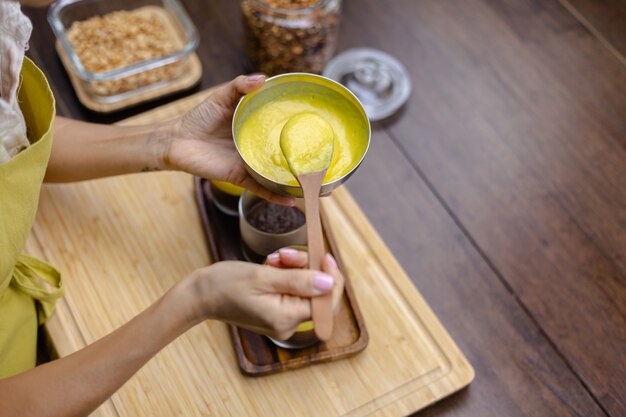 Mujer caucásica en la cocina hace budines de chía con mermelada de mango. Postre elaborado con leche de almendras, semillas de chía, cacao, mermelada de mango y granola.