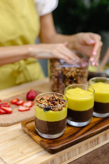 Mujer caucásica en la cocina hace budines de chía con mermelada de mango. Postre elaborado con leche de almendras, semillas de chía, cacao, mermelada de mango y granola.