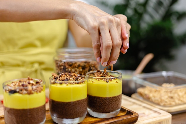 Mujer caucásica en la cocina hace budines de chía con mermelada de mango. Postre elaborado con leche de almendras, semillas de chía, cacao, mermelada de mango y granola.