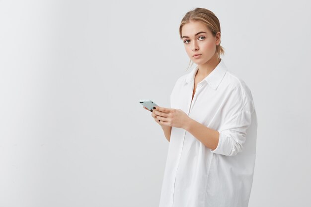 Mujer caucásica confiada con el pelo rubio que lleva el mensaje de mecanografía de la camisa blanca en el teléfono inteligente. Retrato de mujer de negocios seria posando, sosteniendo el teléfono celular en sus manos.