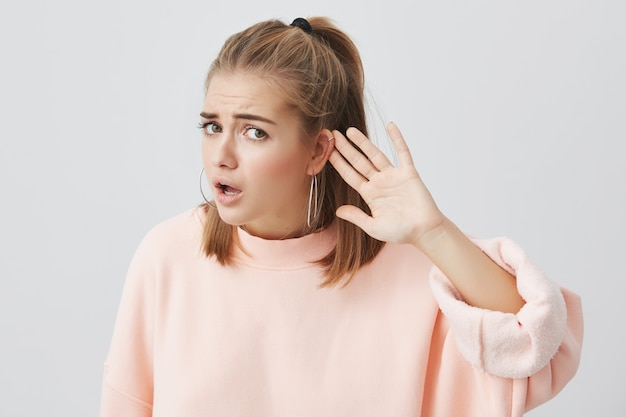 Mujer caucásica desorientada en suéter rosa que mira con expresión confundida y perpleja, frunciendo el ceño, sosteniendo la mano detrás de la oreja como si no escuchara lo que se le dice que haga, y ferviente para hacerlo