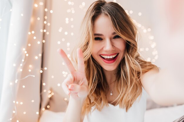 Foto gratuita mujer caucásica despreocupada con maquillaje ligero posando con el signo de la paz. filmación en interiores de la maravillosa chica rizada haciendo selfie en su habitación.