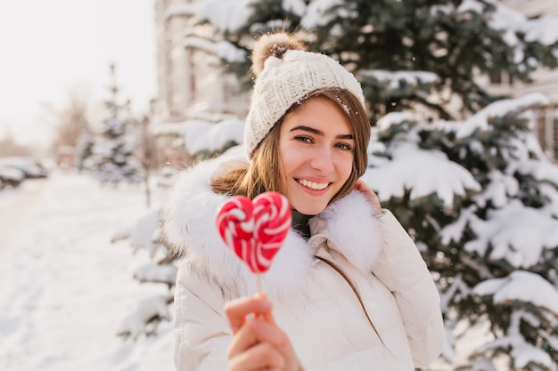 Mujer caucásica entusiasta con piruleta de corazón durante la sesión de fotos de invierno. Alegre mujer lleva gorro de punto y bata blanca posando con caramelos mientras trabaja en el parque nevado