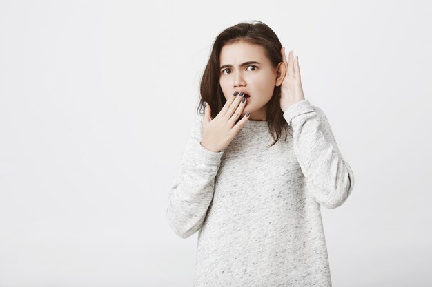 mujer caucásica con expresión sorprendida, cubriendo su boca y sosteniendo la mano cerca de la oreja para escuchar chismes