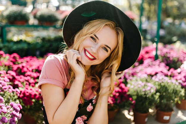 Mujer caucásica extática escalofriante en invernadero en la mañana. Mujer joven inspirada en sombrero negro posando con flores rosas