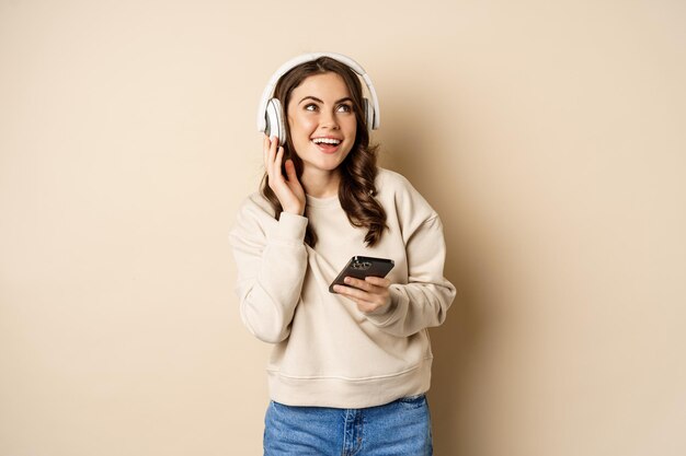 Mujer caucásica feliz escuchando música en auriculares en la aplicación de teléfono inteligente bailando y divirtiéndose standin...