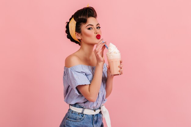 Mujer caucásica glamorosa bebiendo batido. Disparo de estudio de moda chica morena posando con cóctel sobre fondo rosa.