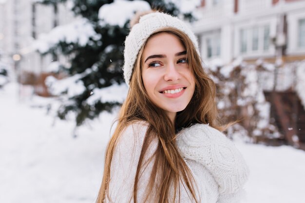 Mujer caucásica inspirada en sombrero de lana mirando a otro lado con una sonrisa mientras posa en la mañana de invierno. Retrato de primer plano de fascinante modelo femenino de pelo largo en suéter blanco suave de pie en el patio cubierto de nieve.