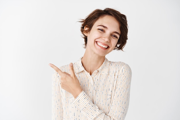 Mujer caucásica joven con belleza natural sin maquillaje sonriendo dientes blancos y señalando con el dedo a la izquierda en el logotipo de pie sobre el fondo del estudio