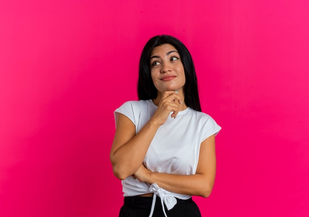 Mujer caucásica joven complacida sostiene la barbilla mirando al lado