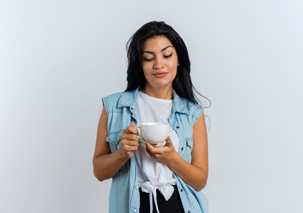 Mujer caucásica joven complacida sostiene y mira la taza