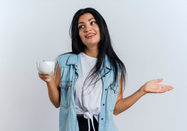 Mujer caucásica joven complacida sostiene la taza y sostiene la mano abierta mirando al lado