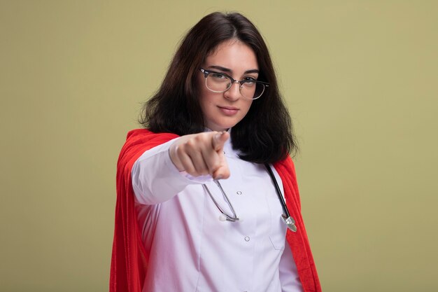 Mujer caucásica joven confiada del superhéroe en capa roja que lleva el uniforme del doctor y el estetoscopio con los vidrios que miran y señalan al frente