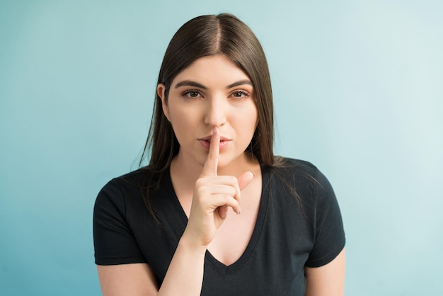 Mujer caucásica joven con el dedo en los labios mientras hace contacto visual contra el fondo liso