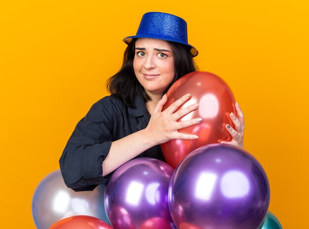 Mujer caucásica joven despistada del partido que lleva el sombrero del partido que se coloca detrás de los globos que sostiene uno que mira al frente aislado en la pared naranja