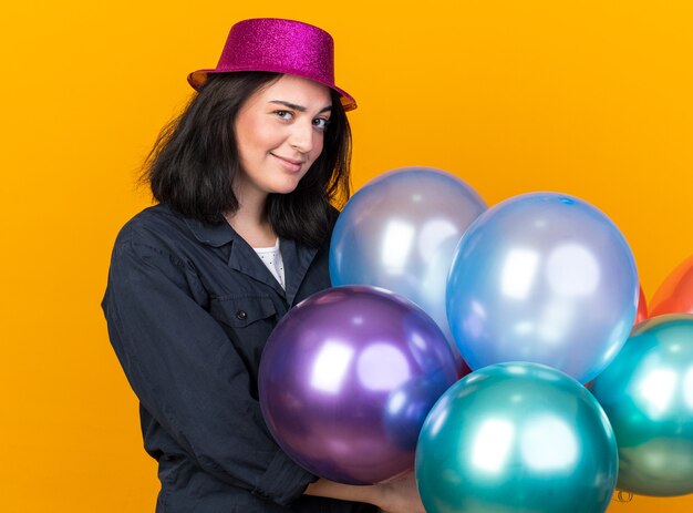 Mujer caucásica joven dudosa del partido que lleva el sombrero del partido que se coloca en vista de perfil que sostiene los globos que miran al frente aislado en la pared naranja