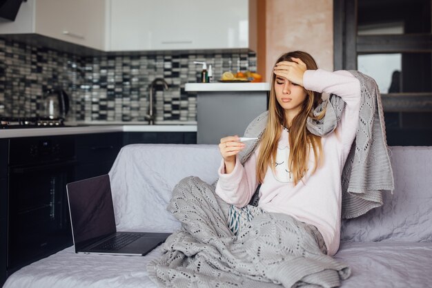 Mujer caucásica joven enferma en ropa de casa gris sentada en la cama con trabajo portátil