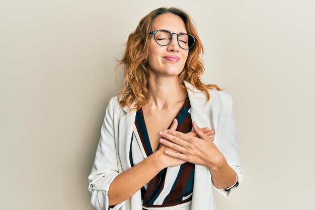 Mujer caucásica joven con estilo de negocios y gafas sonriendo con las manos en el pecho con los ojos cerrados y un gesto agradecido en el concepto de salud facial