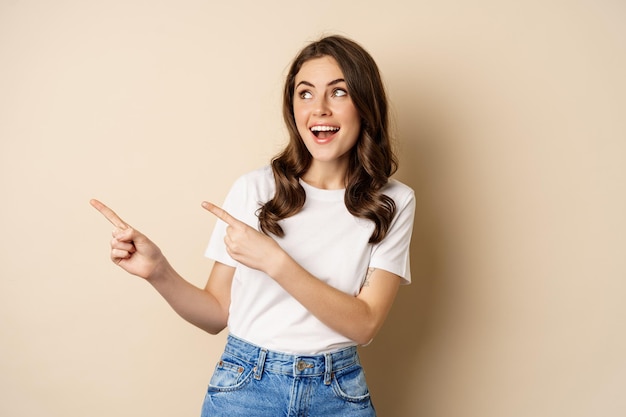 Mujer caucásica joven con estilo sonriendo, señalando con el dedo a la izquierda, mostrando publicidad, oferta promocional, de pie contra un fondo beige.