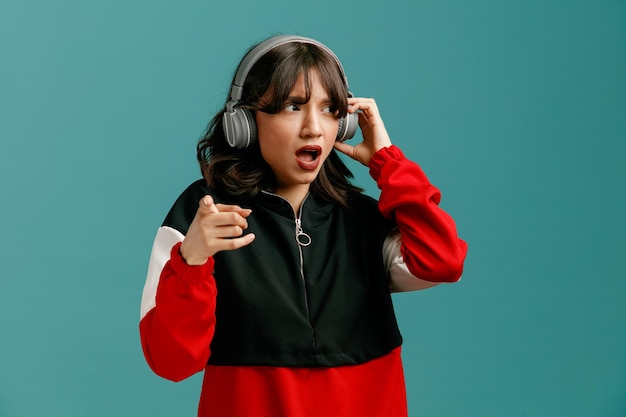 Mujer caucásica joven frunciendo el ceño usando y agarrando auriculares mirando al lado apuntando a la cámara aislada en el fondo azul