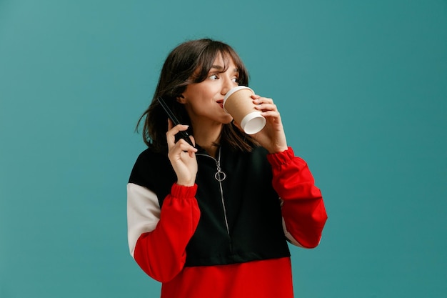 Mujer caucásica joven impresionada hablando por teléfono mirando al lado bebiendo café de una taza de café de papel aislada en el fondo azul