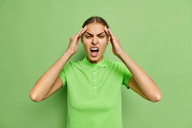 La mujer caucásica joven irritada sufre de un dolor de cabeza terrible mantiene las manos en las sienes mantiene la boca abierta vestida casualmente aislada sobre la pared verde