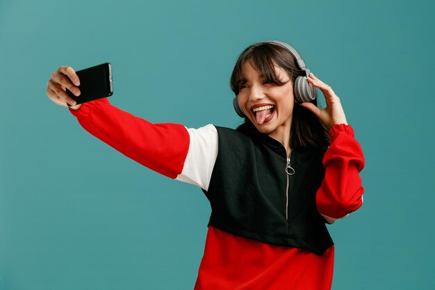 Mujer caucásica joven juguetona usando y agarrando auriculares estirando el teléfono móvil mostrando la lengua tomando selfie aislado sobre fondo azul