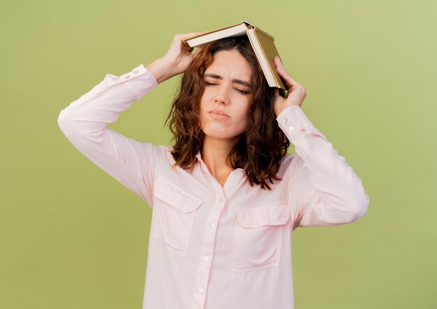 Mujer caucásica joven molesta sostiene el libro sobre la cabeza aislado sobre fondo verde con espacio de copia