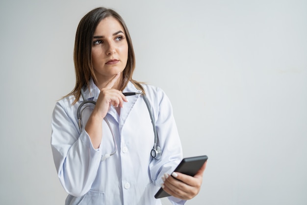 Mujer caucásica joven pensativa en la capa del laboratorio que sostiene el teléfono