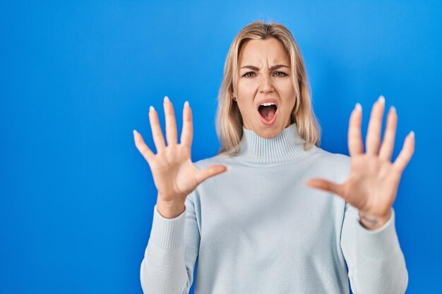 Mujer caucásica joven de pie sobre fondo azul asustada y aterrorizada con expresión de miedo detener el gesto con las manos gritando en concepto de pánico de choque