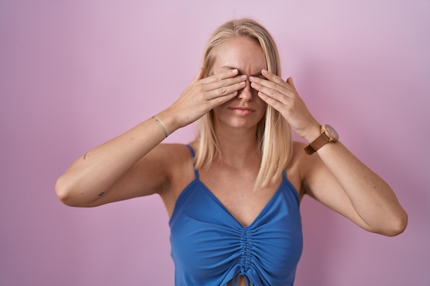 Mujer caucásica joven de pie sobre fondo rosa frotándose los ojos por fatiga y dolor de cabeza, expresión soñolienta y cansada. problema de visión