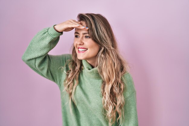 Mujer caucásica joven de pie sobre fondo rosa muy feliz y sonriente mirando lejos con la mano sobre la cabeza. concepto de búsqueda.